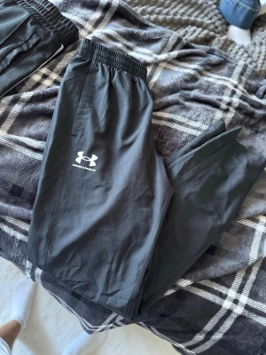 Svarta träningsbyxor Under Armour - Snygga svarta träningsbyxor från Under Armour med elastisk midja och diskret logga på benet. Tillverkade i lätt och snabbtorkande material, perfekta för gymmet eller löprundan. Klassisk passform och stilren design.