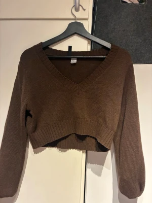 Brun croppad stickad tröja - Säljer en brun croppad stickad tröja med v-ringning och långa ärmar. Tröjan har bred ribbstickad kant vid halsringning, ärmslut och nederkant. Perfekt för lager på lager och en avslappnad stil.