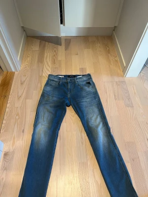 Blå jeans från Replay - Snygga blå jeans från Replay med klassisk femficksdesign. Jeansen har raka ben och normal midja. Perfekta för en avslappnad och trendig look. Jeansen är i nyskick. De svarta trådarna är från en prislapp som jag ej har orkat dra bort för bältet täcker det ändå. 