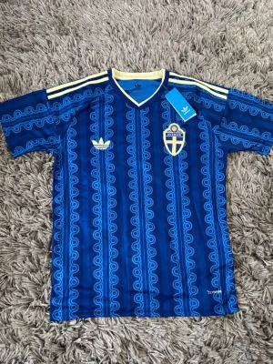 Blå Sverige tröja från Adidas - Säljer en blå fotbollströja från Adidas med Sveriges landslagsemblem på bröstet. Tröjan har gula detaljer, klassiska Adidas-ränder på axlarna och ett retroinspirerat mönster. Tillverkad i lätt och ventilerande material, perfekt för träning eller match.