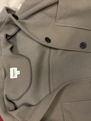 Beige kappa från Kappahl, S - Stilren beige kappa från Kappahl i storlek S. Kappan har klassisk krage, svarta knappar och stora fickor framtill. Materialet är mjukt och känns lite ulligt, perfekt för kyligare dagar. Enkel och tidlös design som passar till det mesta.