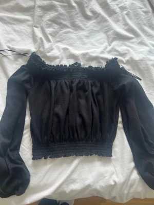 Svart offshoulder blus med volang - Supersöt svart offshoulder blus med långa puffärmar och volangdetaljer vid halsringningen. Smockad midja och ärmslut ger en snygg passform. Perfekt att styla med jeans eller kjol för en trendig look.