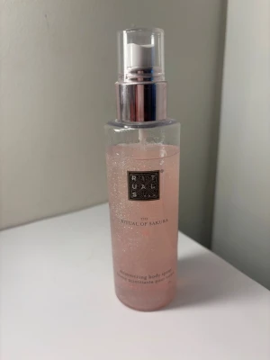 Rituals The Ritual of Sakura Body Spray - Shimmering body spray från Rituals, The Ritual of Sakura. Kommer i en genomskinlig plastflaska med rosa, skimrande vätska och silvrig spraypump. Doften är inspirerad av körsbärsblom och ger huden en lätt glittrande finish. Lite använd vilket syns på bild