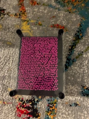 Lila pin art leksak i plast - En klassisk pin art leksak med lila plastpinnar och genomskinlig ram. Perfekt för att skapa 3D-avtryck av händer, ansikten eller andra föremål. Leksaken har en rektangulär form och svarta hörn i plast.