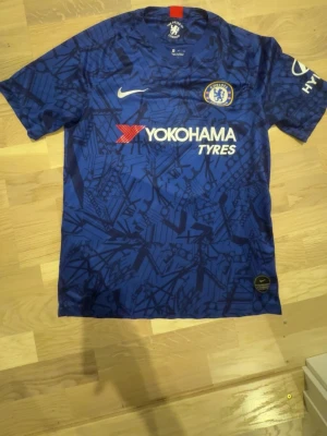 Chelsea matchtröja Kante #7 blå - Chelsea FC matchtröja i blått med mörkblått mönster, officiella loggor och sponsortryck. Kante och nummer 7 på ryggen. Korta ärmar, rund hals och sportig passform. Perfekt för fotbollsträning eller att bära på matchdag.
