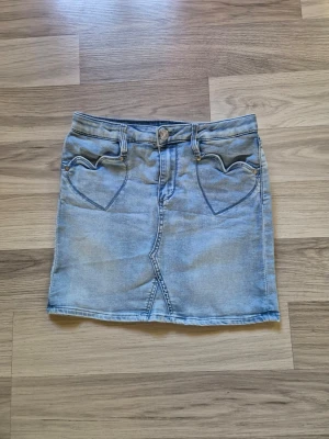Ljusblå jeanskjol med hjärtfickor - Söt ljusblå jeanskjol i kort modell med två hjärtformade fickor fram och klassiska bakfickor. Kjolen har bälteshällor, knapp och dragkedja framtill. Perfekt för en avslappnad och trendig look.