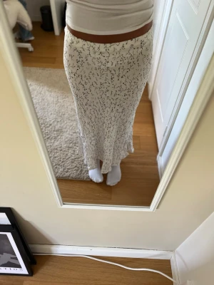 Vit långkjol med svart blommönster - Säljer en vit långkjol med svart blommigt mönster. Kjolen har en rak och något utsvängd passform som sitter snyggt i midjan. Materialet ser ut att vara mjukt och följsamt, perfekt för en trendig och avslappnad look.