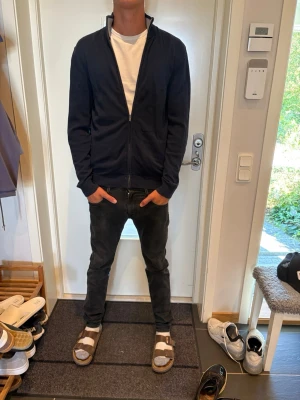 Mörkblå ziptröja med hög krage - Säljer en mörkblå ziptröja med hög krage och dragkedja framtill. Tröjan har en clean och enkel design utan synliga loggor, långa ärmar och ribbade muddar vid ärmslut och nederkant. Perfekt att slänga på sig över en t-shirt för en avslappnad look.