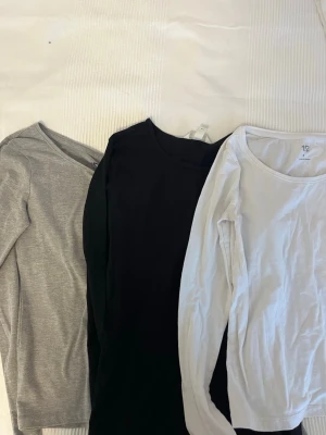 3- pack långärmade tröjor  - En enkel och stilren grå långärmad topp från 157. Tillverkad i en mjuk blandning av bomull, viskos och elastan för skön stretch och passform. Perfekt basplagg med rund halsringning och normal passform. Svart långärmad topp från H&M i storlek S. Vit enkel långärmad topp med rund halsring ifrån Lager 157 i storlek S. 