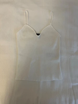 Vit ribbad linnetopp med smala band - Säljer en vit ribbad linnetopp med smala axelband och v-ringad hals. Toppen har en figurnära passform och är perfekt för varma dagar eller som layering-plagg. Enkel och stilren design som passar till det mesta.
