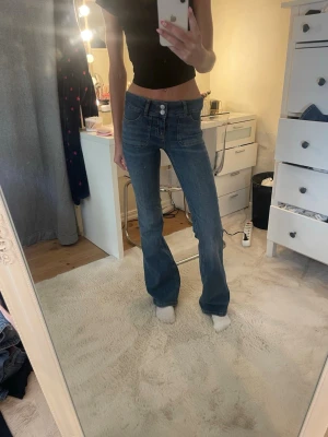 Blå bootcut jeans från Nelly - Säljer ett par blå bootcut jeans från Nelly med låg midja och dubbla knappar framtill. Jeansen har stora framfickor och är tillverkade i ett klassiskt denimtyg. Perfekta för dig som gillar en retroinspirerad look.