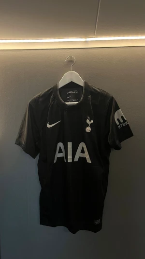 Tottenham Hotspur svart matchtröja - Snygg svart Tottenham Hotspur matchtröja från Nike med klubbens emblem på bröstet och AIA som sponsortryck. Tröjan har korta ärmar, rund hals och diskret rutmönster med texten 'SPURS' i bakgrunden. Perfekt för fotbollsträning eller att bära till vardags.
