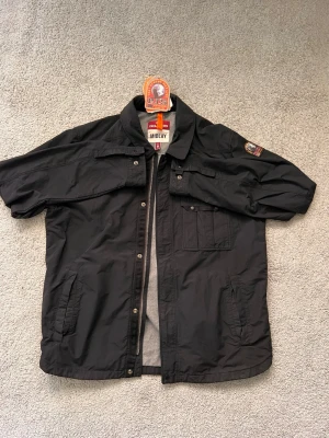 Svart vindjacka från Parajumpers - Säljer en svart vindjacka från Parajumpers, modell MIDLAY. Jackan har flera fickor framtill, kort krage och en patch på ärmen. Tillverkad i lätt polyester med dragkedja och tryckknappar. Perfekt för dig som gillar stilren och funktionell design.