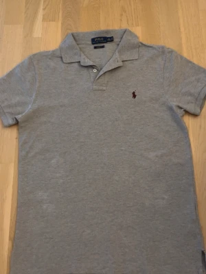 Grå pikétröja från Polo Ralph Lauren - Klassisk grå pikétröja från Polo Ralph Lauren med korta ärmar och liten röd logga broderad på bröstet. Tröjan har krage och knappar vid halsen. Perfekt till jeans eller shorts för en clean look.