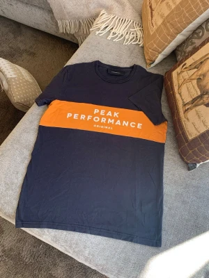 Peak Performance marinblå t-shirt - Snygg marinblå t-shirt från Peak Performance med orange band och vit text över bröstet. Klassisk rund hals och korta ärmar. Tillverkad i mjuk bomull som känns skön mot huden. Perfekt för en avslappnad och sportig stil.