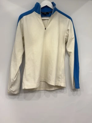 Vit och blå fleece från Everest - Säljer en vit fleece från Everest med blå detaljer längs ärmar och krage. Tröjan har halv dragkedja framtill och hög krage för extra värme. Perfekt för kyliga dagar och enkel att matcha med andra plagg.