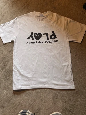 Vit Comme des Garçons PLAY t-shirt - Säljer en vit t-shirt från Comme des Garçons PLAY med det ikoniska hjärtloggan och texten PLAY tryckt i svart på bröstet. Klassisk passform och rund halsringning. Perfekt för dig som gillar streetwear och stilrena plagg.