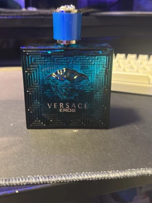 Versace Eros parfym blå flaska - Versace Eros parfym i en snygg, fyrkantig blå glasflaska med silverdetaljer och ett coolt Medusa-motiv på framsidan. Flaskan har en blå och silverfärgad kork som ger en lyxig känsla. Perfekt för dig som gillar stilrena och exklusiva dofter.
