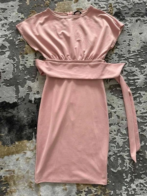 Ljusrosa omlottklänning med bälte - Snygg ljusrosa klänning med omlottdetalj och brett midjebälte. Klänningen har korta ärmar och en rak, kort siluett. Perfekt för dig som gillar en feminin och enkel stil.