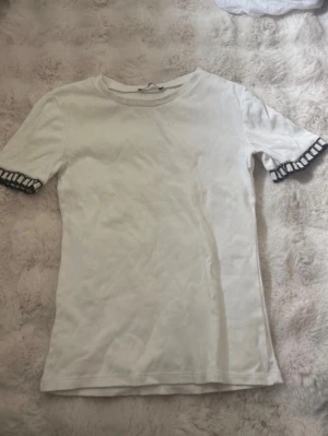Vit t-shirt med volangärm från Zara - Säljer en vit t-shirt från Zara med rund hals och korta ärmar. Ärmsluten har en snygg volangdetalj med svartvitt mönster som ger en unik touch. Tillverkad i mjukt bomullsmaterial som känns skönt mot huden. Perfekt för dig som gillar stilrena plagg med en twist.