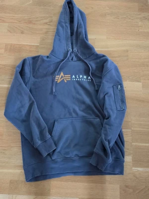 Grå hoodie Alpha Industries - Snygg grå hoodie från Alpha Industries med stor huva, dragsko och känguruficka. Framtill finns en vit och gul logga, och på ena ärmen sitter en cool ficka med dragkedja och Alpha-tag. Tillverkad i mjuk bomullsmix.