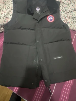 Svart väst från Canada Goose - Säljer en svart dunväst från Canada Goose med klassisk logga på bröstet och diskret varumärkesmärke på fickan. Västen har hög krage, tryckknappar och dragkedja framtill. Perfekt för lager-på-lager och håller dig varm under kyliga dagar. Passar s-L