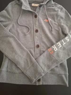 Grå hoodie från Hollister med knappar - Grå hoodie från Hollister med knäppning framtill och dragsko i huvan. Broderad logga på bröstet och tryck på ena ärmen med texten 'Hollister 22'. Mjuk bomullsblandning och två fickor framtill. Perfekt för en avslappnad stil.