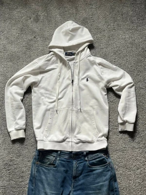 Vit hoodie från Polo Ralph Lauren - Vit hoodie med dragkedja från Polo Ralph Lauren. Klassisk design med huva, dragsnören och två fickor framtill. Broderad marinblå logga på bröstet. Storlek Medium mer som Small!