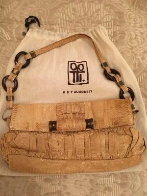 Beige handväska i ormskinnslook - Unik beige handväska från R & Y Augousti i ormskinnslook. Väskan har ett dekorativt handtag med stora ringar i mörkbrunt och beige, samt ett lock med snygg detalj framtill. Perfekt accessoar för dig som vill sticka ut.