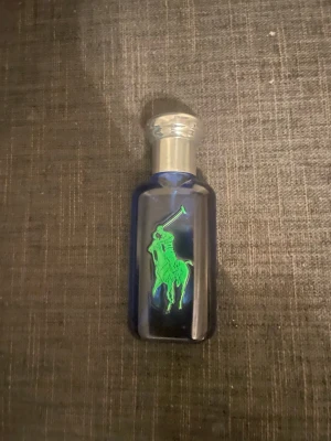 Blå parfymflaska med grön logga - Snygg parfymflaska i blått glas med en stor grön logga av en ryttare på framsidan och en gul siffra 1 på baksidan. Flaskan har ett silverfärgat lock och en modern, sportig design som sticker ut i badrumshyllan.