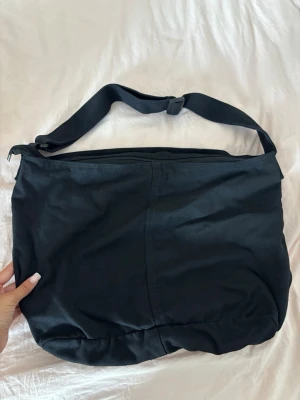 Svart väska  - Enkel och rymlig svart shoppingväska i canvas med justerbar axelrem. Väskan har en mjuk, slouchy form och stängs med dragkedja upptill. Perfekt för att bära med sig allt man behöver under dagen.