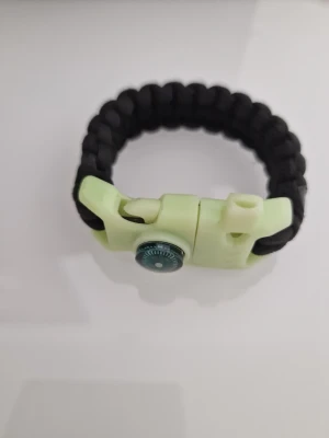 Svart paracordarmband med kompass - Snyggt armband i svart paracord med ett spänne i ljusgrön plast och inbyggd liten kompass, visselpipa, flinta samt ska lysa lite i mörkret. Perfekt för dig som gillar äventyr och vill ha en cool accessoar med praktisk funktion. 