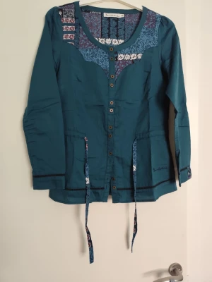 Turkos blus med blommiga detaljer - Turkos blus med lång ärm, knappar framtill och rund hals. Blommiga mönster i blått, vitt och vinrött vid axlar och halsringning samt matchande knytband i midjan. Materialet ser ut att vara tunn bomull eller viskos.