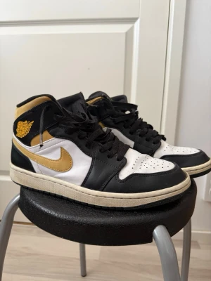 Air Jordan 1 mid white pollen - Snygga sneakers i svart och vitt med guldiga detaljer och swoosh. Skorna har klassisk snörning, perforerad tåbox och en kontrasterande vit sula. Materialet är en mix av läder och textil. Perfekta för dig som vill sticka ut med stilrena färgkombinationer.