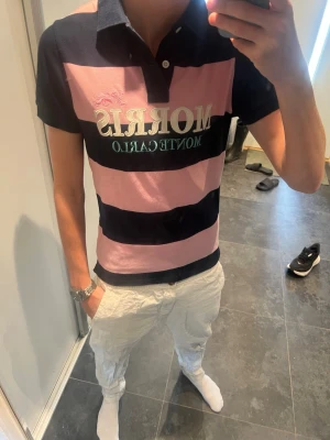 Morrus pike - Säljer en snygg pikétröja från Morris med breda marinblå och rosa ränder. Tröjan har klassisk krage, korta ärmar och stor logga med texten 'Morris Monte Carlo' på bröstet. Perfekt för en sportig och stilren look.
