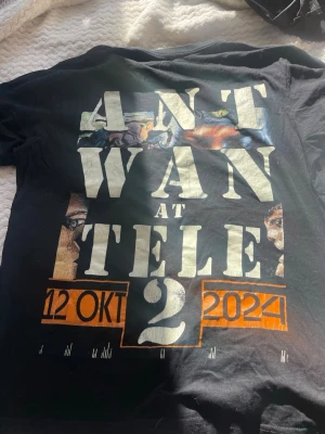 Svart t-shirt med Ant Wan-tryck - Cool svart t-shirt med stort tryck på framsidan som visar texten 'Ant Wan at Tele2', datum och grafiska detaljer i vitt och orange. Baksidan har tryck med information om eventet. Perfekt för dig som gillar streetwear och musikmerch.