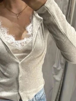 Beige omlottopp med spetsdetaljer - Säljer en beige omlottopp med långa ärmar och fina spetsdetaljer vid halsringningen. Toppen har en v-ringad design och knappar framtill. Perfekt för dig som gillar en feminin och stilren look.