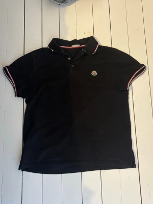 Moncler pikétröja - Klassisk svart pikétröja från Moncler med korta ärmar och vit-röda detaljer på krage och ärmslut. Tröjan har knappslå framtill och Moncler-logga broderad på bröstet. Perfekt för en stilren och sportig look.