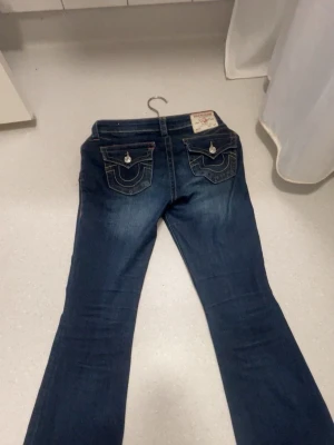 Mörblå true religion jeans w27 - Säljer ett par mörkblå bootcut jeans med snygga fickdetaljer och kontrastsömmar. Jeansen har två bakfickor med lock och knapp, samt klassisk femficksdesign. Materialet är denim med lätt stretch för skön passform.
