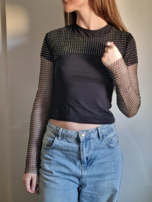 Svart mesh topp med nätärmar glitter - Snygg svart mesh topp med långa nätärmar och croppad passform. Toppen har små glittriga detaljer över hela tyget och är perfekt för dig som vill sticka ut. Materialet är tunt och stretchigt syntet. Passar till många olika stilar.