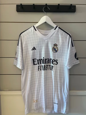 Real Madrid matchtröja Bellingham - Säljer en vit Real Madrid matchtröja från Adidas med Bellingham och nummer 5 på ryggen. Tröjan har svarta detaljer, klubbmärke på bröstet och sponsortryck. Tillverkad i lätt och ventilerande material, perfekt för fotbollsträning eller match.