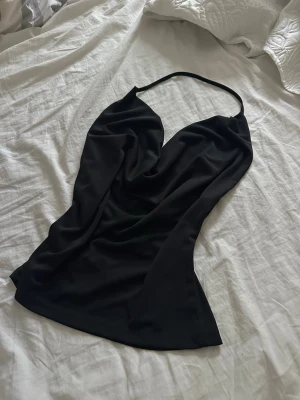 Svart axelbandslös topp - Svart axelbandslös topp med halterneck och draperad front. Den har öppen rygg och är tillverkad i ett fint, följsamt syntetmaterial som ger en snygg siluett.