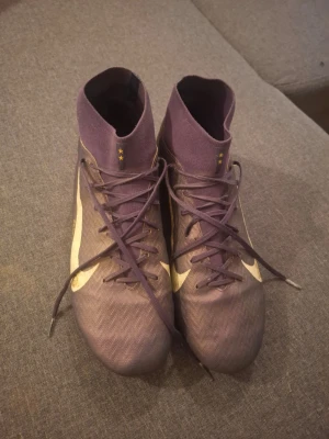 Lila Nike fotbollsskor med högt skaft - Säljer ett par Mbappe x nike fotbollsskor från Nike med högt skaft och vita detaljer. Skorna har snörning framtill och är tillverkade i ett syntetmaterial som ger bra bollkontroll. Perfekta för dig som vill sticka ut på planen med stilrena och sportiga skor. Säljes pga för små