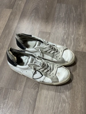 Vita och svarta låga sneakers - Säljer ett par vita sneakers med svarta detaljer och svart häl. Skorna har vita snören och en klassisk låg siluett. Yttermaterialet är troligtvis läder eller syntet och sulan är svart med mönstrat grepp. Perfekta för en avslappnad streetstil. Lite sönder från insidan men utsidan är de perfekt, storlek 42
