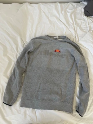 Grå sweatshirt från Ellesse - Snygg grå sweatshirt från Ellesse med klassisk logga i grått och färgdetaljer på bröstet. Tröjan har rund halsringning, långa ärmar och svarta detaljer vid ärmsluten. Perfekt för en avslappnad och sportig stil.