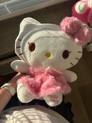 Hello Kitty mjukisdjur med rosa klänning - 35x30 cm stor Söt Hello Kitty mjukisdjur i vitt plyschmaterial med rosa fluffig klänning och stor rosa rosett på huvudet. Broderad text 'Hello Kitty' på ena tassen och bruna broderade ögon. Perfekt som present eller dekoration.