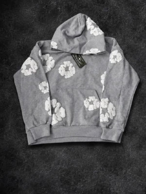 Grå hoodie med vita blommor - Snygg grå hoodie med vita blommönster över hela tröjan. Hoodien har en klassisk känguruficka framtill och en rymlig huva. Perfekt för dig som vill ha en chill och trendig stil. Tillverkad i mjukt material för extra komfort.