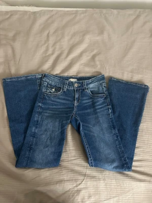Blå bootcut jeans med broderade fickor - Säljer ett par blå bootcut jeans med snygga broderade detaljer på bakfickorna och kontrastsömmar. Jeansen har klassisk femficksmodell, normal midja och är tillverkade i denim med lätt tvättad look. Perfekta för dig som gillar en avslappnad men trendig stil. Använt typ en gång.