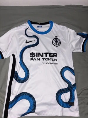 Inter Milan vit matchtröja med orm - Snygg vit Inter Milan matchtröja från Nike med blå och svart orm-mönster längs framsidan. Tröjan har klubbmärke och Nike-logga på bröstet, samt sponsortryck. Rund hals med blå kant och svarta detaljer på ärmarna. Perfekt för fotbollsfans.