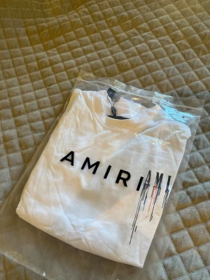 Vit AMIRI t-shirt med färgstänk - Säljer en vit t-shirt från AMIRI med svart logotyp framtill och coolt färgstänksmönster i svart, blått och rött både fram och bak. T-shirten har korta ärmar och är gjord i mjuk bomull. Perfekt för dig som gillar streetwear vibes.